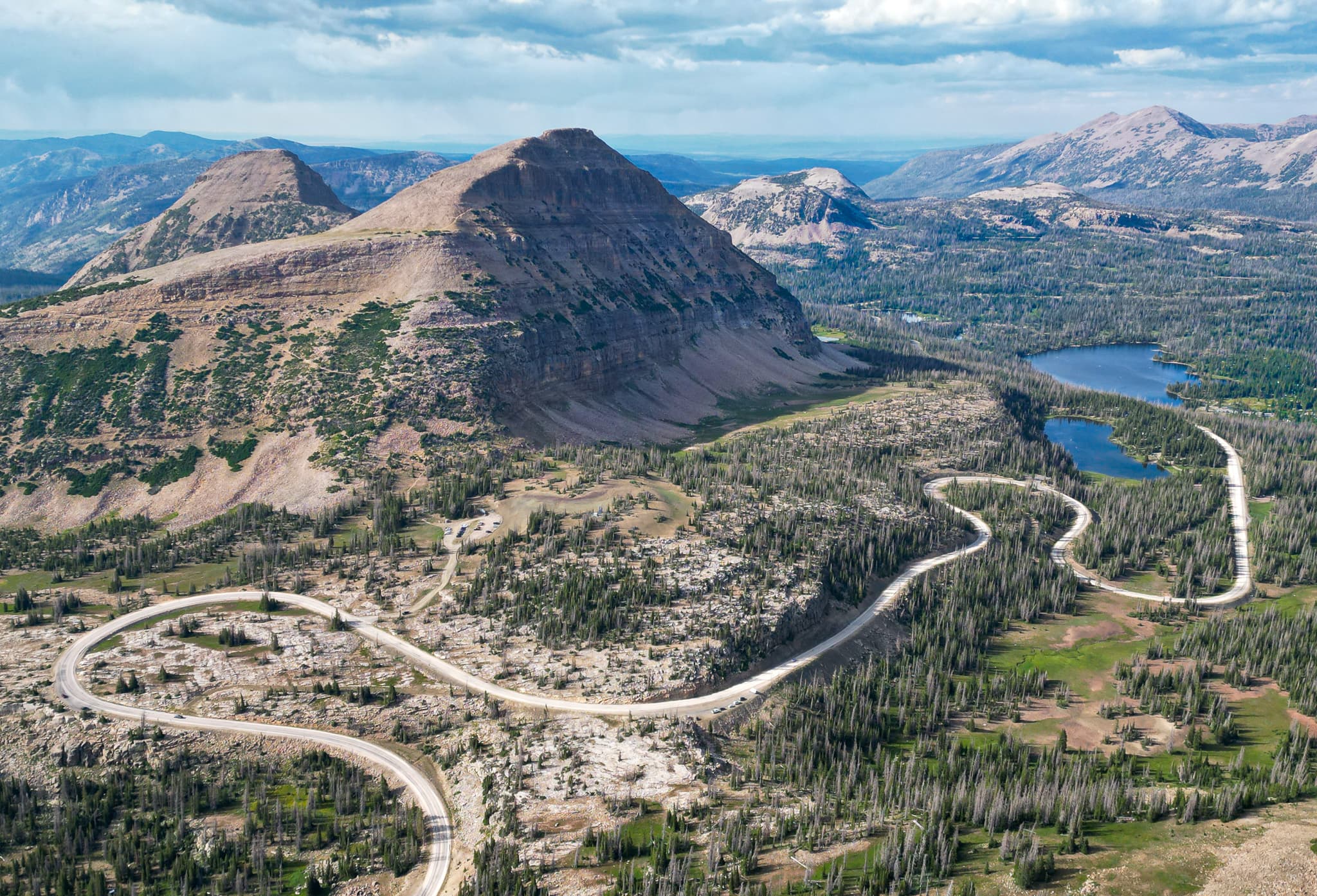 A winding road traverses a mountainous landscape with a prominent peak and a nearby lake, surrounded by lush greenery and distant mountain ranges under a partly cloudy sky