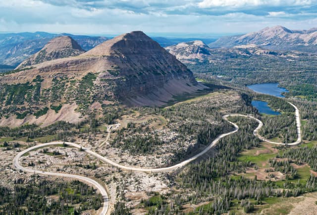 A winding road traverses a mountainous landscape with a prominent peak and a nearby lake, surrounded by lush greenery and distant mountain ranges under a partly cloudy sky