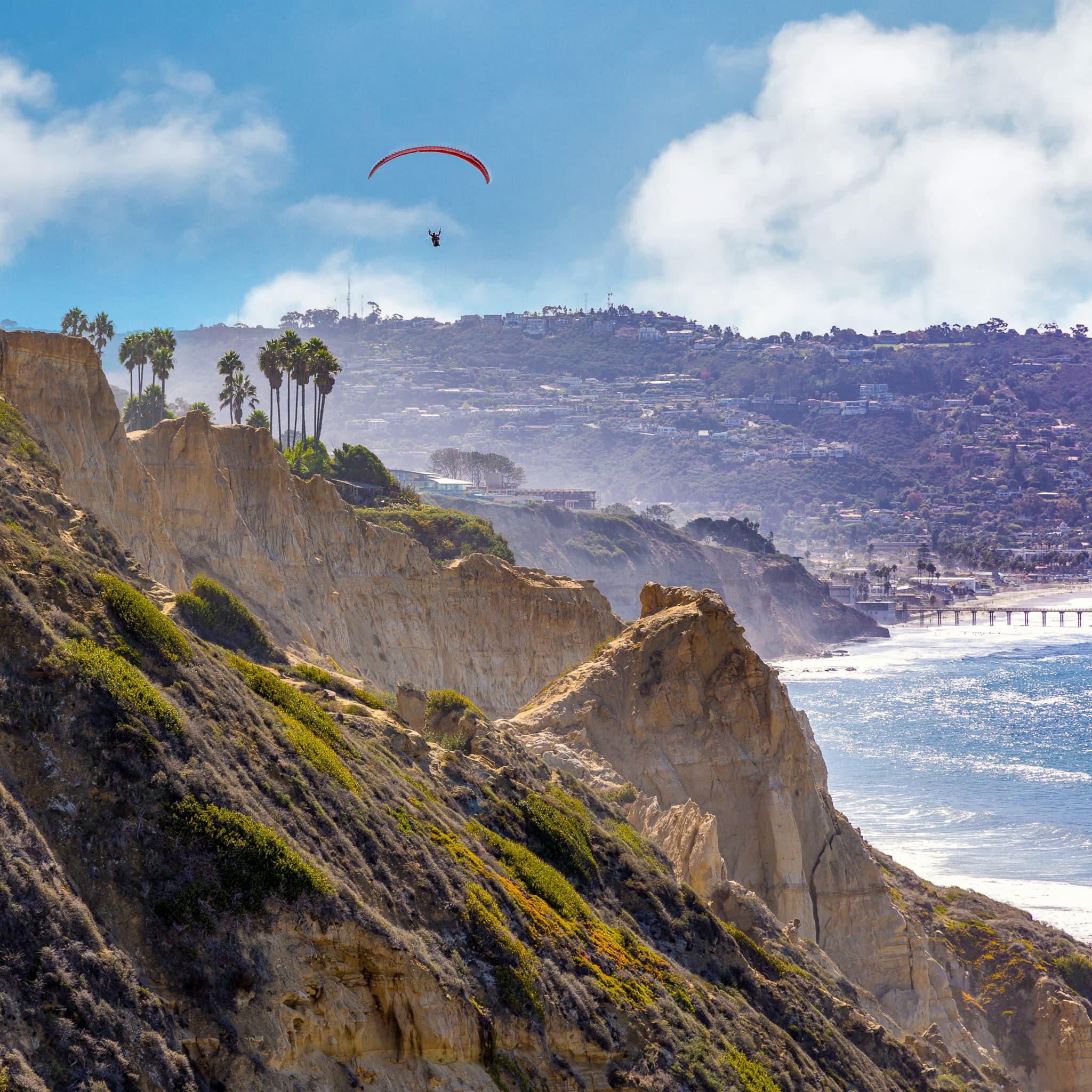 A coastal cliffside with a paraglider in the sky, overlooking the ocean and a distant pier