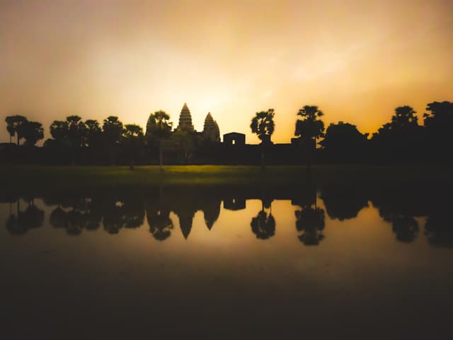 A silhouette of a temple complex with trees is reflected in a calm body of water during a golden sunset