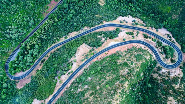 A winding road curves through a lush, green landscape with dense vegetation and patches of exposed earth