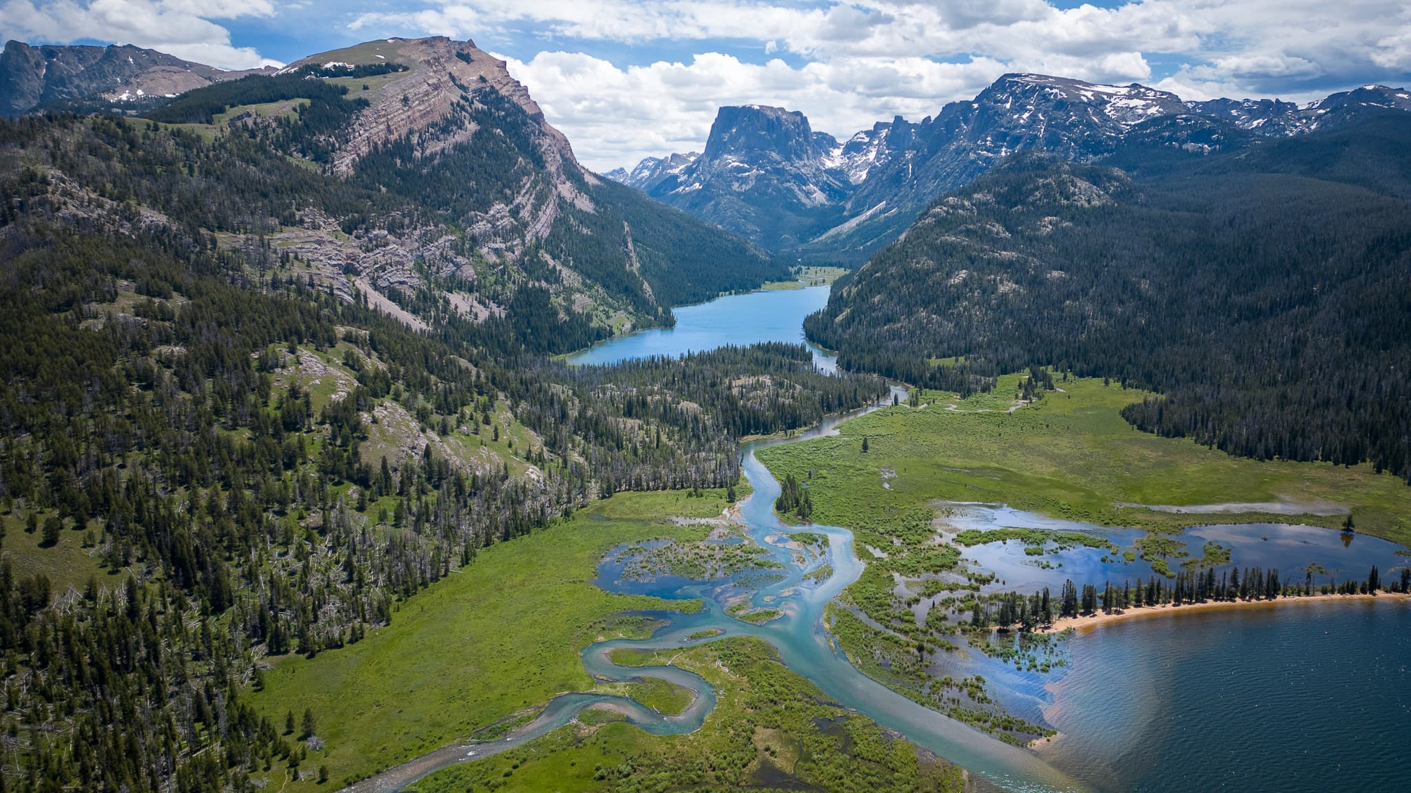 A scenic landscape featuring a winding river flowing through a lush green valley, surrounded by forested mountains and a lake, under a partly cloudy sky