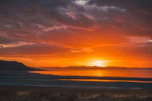 A stunning sunset over a calm body of water, with vibrant orange and red hues in the sky, silhouetted mountains in the background, and a dark shoreline in the foreground