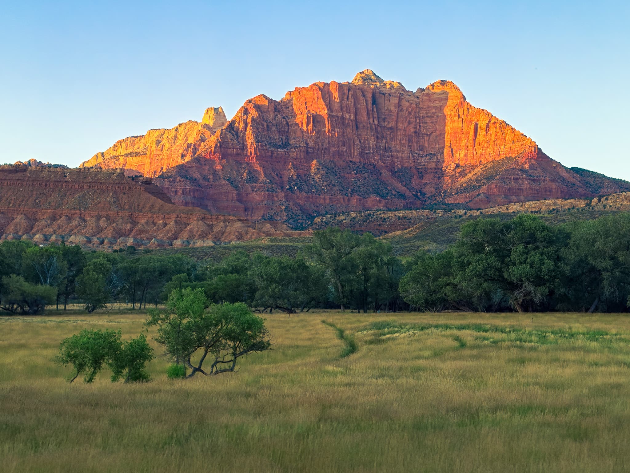 A majestic mountain range bathed in warm sunlight, with a foreground of green fields and scattered trees